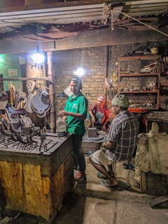Several people are working in a dimly lit workshop. A man in a green shirt is standing near a table with various metal tools, another sits on a chair in plaid clothing, and a person in red takes a break sitting on a block in the background. The room contains worn brick walls, shelves cluttered with tools, and dim overhead lighting.