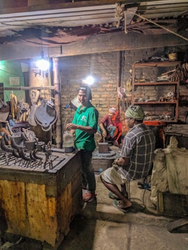 Several people are working in a dimly lit workshop. A man in a green shirt is standing near a table with various metal tools, another sits on a chair in plaid clothing, and a person in red takes a break sitting on a block in the background. The room contains worn brick walls, shelves cluttered with tools, and dim overhead lighting.