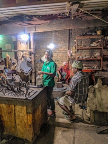 Several people are working in a dimly lit workshop. A man in a green shirt is standing near a table with various metal tools, another sits on a chair in plaid clothing, and a person in red takes a break sitting on a block in the background. The room contains worn brick walls, shelves cluttered with tools, and dim overhead lighting.