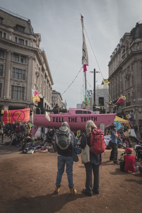 A protest scene in an urban setting featuring a large pink boat with the words 'Tell The Truth' written on the side. The boat is surrounded by a crowd of people, some of whom are carrying backpacks, while others are sitting or standing around. Colorful flags and banners with various symbols and messages are displayed around the area. The scene is set between two large, ornate buildings under a cloudy sky.