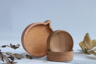 Wooden containers with a simple, elegant design are placed in the center; one resembles an apple shape. Accompanied by dried leaves on a light wooden surface, all set against a soft blue background.