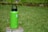 A vibrant green water bottle sits on a tree stump, standing out against the lush green grass in the background. The bottle's lid is black with a handle, and there's a white logo on its front.