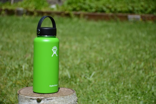 A vibrant green water bottle sits on a tree stump, standing out against the lush green grass in the background. The bottle's lid is black with a handle, and there's a white logo on its front.