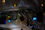 Stage lighting setup featuring blue spotlights attached to metal trusses under a tent-like fabric ceiling. A circular emblem with a green laurel wreath is visible among the equipment.