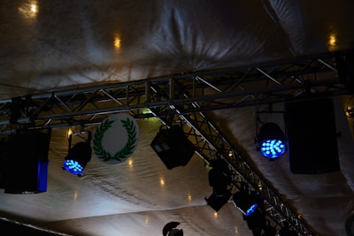 Stage lighting setup featuring blue spotlights attached to metal trusses under a tent-like fabric ceiling. A circular emblem with a green laurel wreath is visible among the equipment.