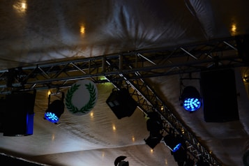 Stage lighting setup featuring blue spotlights attached to metal trusses under a tent-like fabric ceiling. A circular emblem with a green laurel wreath is visible among the equipment.
