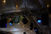 Stage lighting setup featuring blue spotlights attached to metal trusses under a tent-like fabric ceiling. A circular emblem with a green laurel wreath is visible among the equipment.
