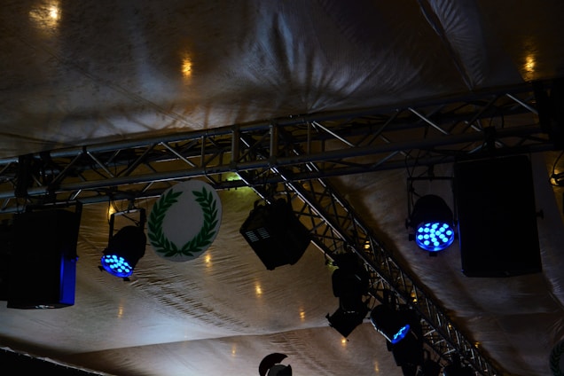 Stage lighting setup featuring blue spotlights attached to metal trusses under a tent-like fabric ceiling. A circular emblem with a green laurel wreath is visible among the equipment.