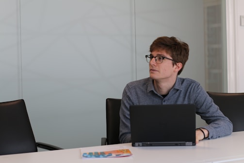 A person with glasses, seated at a conference table, looks to the side while using a laptop. The setting is an office space with a minimalist design, featuring a brochure on the table.