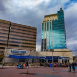 Two tall commercial buildings with external signs for Wells Fargo and Zions Bank alongside a restaurant named Lucky Fins featuring a blue awning. Tree-lined paved area in front with people sitting at tables and a rack with parked bicycles.