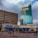 Two tall commercial buildings with external signs for Wells Fargo and Zions Bank alongside a restaurant named Lucky Fins featuring a blue awning. Tree-lined paved area in front with people sitting at tables and a rack with parked bicycles.