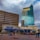 Two tall commercial buildings with external signs for Wells Fargo and Zions Bank alongside a restaurant named Lucky Fins featuring a blue awning. Tree-lined paved area in front with people sitting at tables and a rack with parked bicycles.