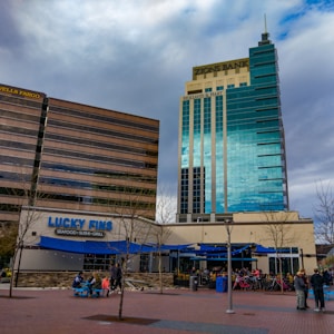 Two tall commercial buildings with external signs for Wells Fargo and Zions Bank alongside a restaurant named Lucky Fins featuring a blue awning. Tree-lined paved area in front with people sitting at tables and a rack with parked bicycles.