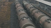 A series of large, cylindrical freight train cars lined up on tracks beside a train platform. The cars appear weathered, with a muted brown color. The surrounding area includes gravel tracks and sparse vegetation alongside the tracks.