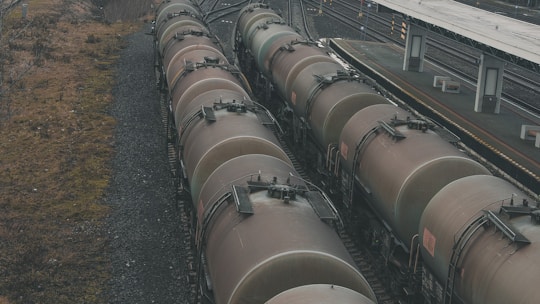 A series of large, cylindrical freight train cars lined up on tracks beside a train platform. The cars appear weathered, with a muted brown color. The surrounding area includes gravel tracks and sparse vegetation alongside the tracks.