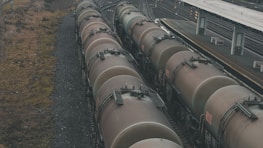 A series of large, cylindrical freight train cars lined up on tracks beside a train platform. The cars appear weathered, with a muted brown color. The surrounding area includes gravel tracks and sparse vegetation alongside the tracks.