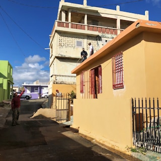 A neighborhood scene with colorful buildings, including a beige house with red window grills and a green building in the background. Two individuals are on the rooftop of the beige house, seemingly engaged in painting or construction work, while another person stands on the street holding up what appears to be an object or device. The street is narrow and paved, with a metal fence lining the beige house. The sunlight casts strong shadows, indicating a bright and sunny day with clear blue skies and a few fluffy clouds.