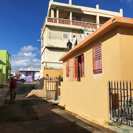 A neighborhood scene with colorful buildings, including a beige house with red window grills and a green building in the background. Two individuals are on the rooftop of the beige house, seemingly engaged in painting or construction work, while another person stands on the street holding up what appears to be an object or device. The street is narrow and paved, with a metal fence lining the beige house. The sunlight casts strong shadows, indicating a bright and sunny day with clear blue skies and a few fluffy clouds.