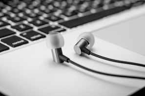 Close-up of earphones resting on a laptop keyboard. The earphones are metallic with black cables, positioned on the white base of the keyboard. The keyboard features black keys with white lettering.