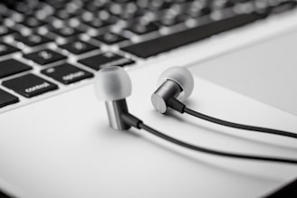 Close-up of earphones resting on a laptop keyboard. The earphones are metallic with black cables, positioned on the white base of the keyboard. The keyboard features black keys with white lettering.