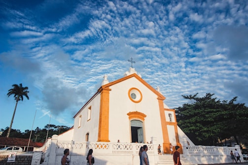 A historical church with white walls and orange accents stands prominently against a vibrant blue sky dotted with clouds. Several people walk and gather near the steps leading to the entrance. A tall palm tree and other greenery can be seen in the background, adding to the serene atmosphere.