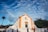 A historical church with white walls and orange accents stands prominently against a vibrant blue sky dotted with clouds. Several people walk and gather near the steps leading to the entrance. A tall palm tree and other greenery can be seen in the background, adding to the serene atmosphere.