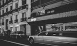 A nighttime urban scene featuring a street lined with parked cars, including a prominent white limousine. The buildings have classic architecture with balconies and decorative molding. Storefronts with illuminated signs add to the urban atmosphere, and a person is standing near the building.