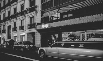 A nighttime urban scene featuring a street lined with parked cars, including a prominent white limousine. The buildings have classic architecture with balconies and decorative molding. Storefronts with illuminated signs add to the urban atmosphere, and a person is standing near the building.