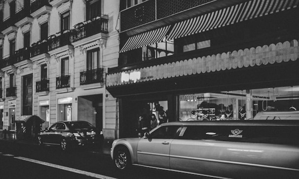 A nighttime urban scene featuring a street lined with parked cars, including a prominent white limousine. The buildings have classic architecture with balconies and decorative molding. Storefronts with illuminated signs add to the urban atmosphere, and a person is standing near the building.