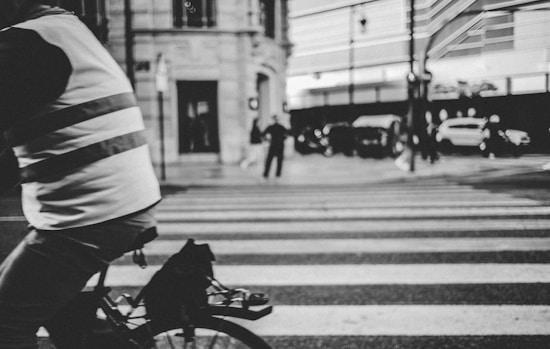 A cyclist wearing a reflective vest rides across a crosswalk in an urban environment. The street is busy with several people and cars in the background, blurred to show motion and activity.