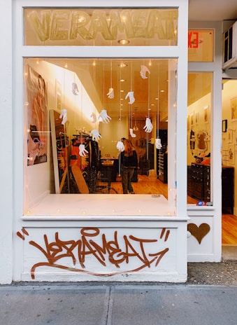 A storefront display with a large window decorated with hanging white gloves. Inside, there is a warm, inviting interior with wooden floors and various items on display. The store name 'VERAMEAT' is visible at the top of the window. Below the window, brown graffiti is painted on the facade. An adjacent door is adorned with a gold heart sticker.