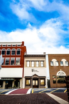 A row of three distinct storefronts set against a backdrop of vivid blue sky with scattered clouds. The central building is marked as a law firm, flanked by a red-brick facade with arches and another with a patterned brick exterior labeled 'Hong Kong Cafe'. A pedestrian crosswalk and brick pavement are visible in the foreground.