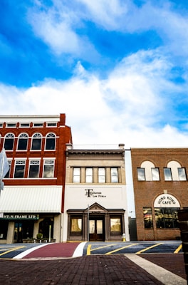 A row of three distinct storefronts set against a backdrop of vivid blue sky with scattered clouds. The central building is marked as a law firm, flanked by a red-brick facade with arches and another with a patterned brick exterior labeled 'Hong Kong Cafe'. A pedestrian crosswalk and brick pavement are visible in the foreground.
