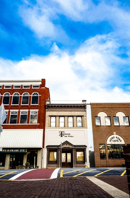 A row of three distinct storefronts set against a backdrop of vivid blue sky with scattered clouds. The central building is marked as a law firm, flanked by a red-brick facade with arches and another with a patterned brick exterior labeled 'Hong Kong Cafe'. A pedestrian crosswalk and brick pavement are visible in the foreground.