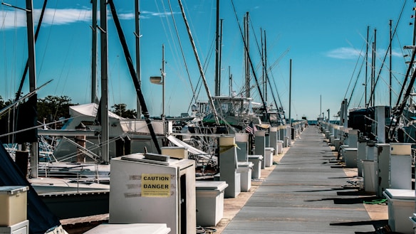 A marina with numerous sailboats and yachts docked along a pier. The wooden pathway is bordered by electrical boxes labeled with cautionary signs. Tall masts of the boats rise against a clear blue sky.