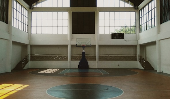 A spacious indoor basketball court with a central hoop and wooden flooring. Large windows allow natural light to stream into the open space, casting shadows on the court. Staircases on either side lead to a balcony with railings, enhancing the airy ambiance.