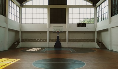 A spacious indoor basketball court with a central hoop and wooden flooring. Large windows allow natural light to stream into the open space, casting shadows on the court. Staircases on either side lead to a balcony with railings, enhancing the airy ambiance.