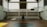 A spacious indoor basketball court with a central hoop and wooden flooring. Large windows allow natural light to stream into the open space, casting shadows on the court. Staircases on either side lead to a balcony with railings, enhancing the airy ambiance.