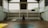 A spacious indoor basketball court with a central hoop and wooden flooring. Large windows allow natural light to stream into the open space, casting shadows on the court. Staircases on either side lead to a balcony with railings, enhancing the airy ambiance.