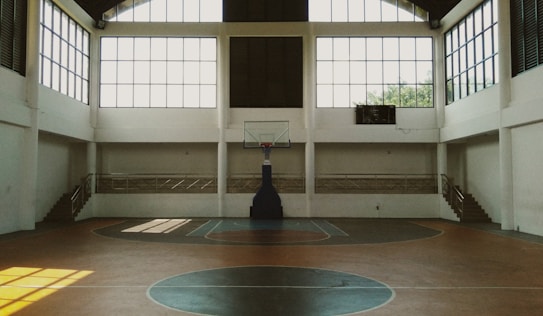 A spacious indoor basketball court with a central hoop and wooden flooring. Large windows allow natural light to stream into the open space, casting shadows on the court. Staircases on either side lead to a balcony with railings, enhancing the airy ambiance.