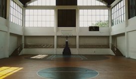A spacious indoor basketball court with a central hoop and wooden flooring. Large windows allow natural light to stream into the open space, casting shadows on the court. Staircases on either side lead to a balcony with railings, enhancing the airy ambiance.