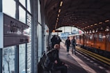 A train station platform with an orange train on the right side and several people walking and waiting. The overhang of the platform has an industrial design with metal beams and soft lighting. A sign on the left indicates the station's name. The atmosphere appears somewhat busy with a few people seated and standing.