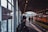 A train station platform with an orange train on the right side and several people walking and waiting. The overhang of the platform has an industrial design with metal beams and soft lighting. A sign on the left indicates the station's name. The atmosphere appears somewhat busy with a few people seated and standing.