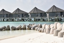 A tranquil beach scene featuring a series of gray wooden bungalows built on stilted platforms over clear turquoise water. The foreground shows a sandy beach with large concrete wave breakers arranged along the shoreline. Seabirds can be seen perched on the wave breakers. The sky is lightly overcast.