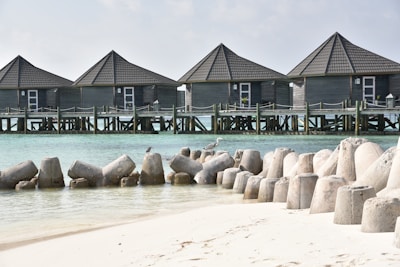 A tranquil beach scene featuring a series of gray wooden bungalows built on stilted platforms over clear turquoise water. The foreground shows a sandy beach with large concrete wave breakers arranged along the shoreline. Seabirds can be seen perched on the wave breakers. The sky is lightly overcast.