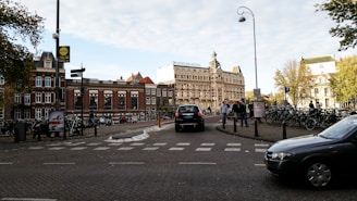 A bustling urban scene featuring a historic brick building with a prominent sign reading 'Amstelkring Hotel'. Numerous bicycles are parked along the street, and pedestrians walk nearby. Several cars are visible on the cobblestone road, which adds to the lively atmosphere. Mature trees with green foliage are present, adding a touch of nature to the cityscape.