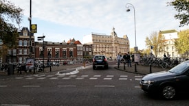 A bustling urban scene featuring a historic brick building with a prominent sign reading 'Amstelkring Hotel'. Numerous bicycles are parked along the street, and pedestrians walk nearby. Several cars are visible on the cobblestone road, which adds to the lively atmosphere. Mature trees with green foliage are present, adding a touch of nature to the cityscape.