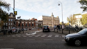 A bustling urban scene featuring a historic brick building with a prominent sign reading 'Amstelkring Hotel'. Numerous bicycles are parked along the street, and pedestrians walk nearby. Several cars are visible on the cobblestone road, which adds to the lively atmosphere. Mature trees with green foliage are present, adding a touch of nature to the cityscape.