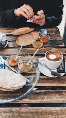 A wooden table set with a breakfast spread that includes slices of toasted bread, small glass bowls of different jams, a cup of cappuccino, and a person opening a small packet. The scene portrays a cozy and casual dining setup.