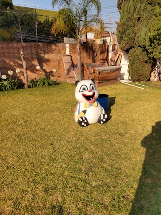 A panda statue with a cheerful expression is sitting on a grassy lawn. It features vibrant colors, including a yellow bow tie and multicolored bands on its body. The background includes a brick wall, some trees, and gardening elements, creating a peaceful outdoor setting with clear sunny weather.